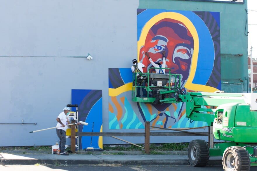 Three people using heavy machinery and paint supplies constructing a colorful portrait mural on the side of a building.